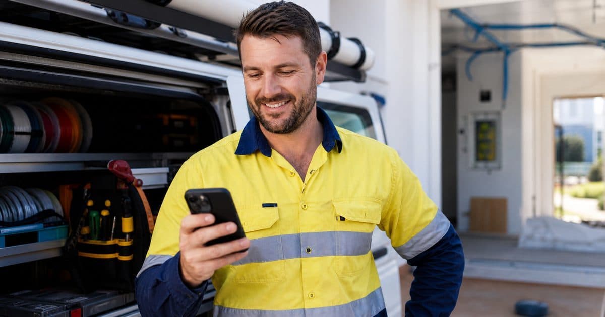 Australian electrician in a hi-vis shirt checking job leads on a smartphone beside a work van